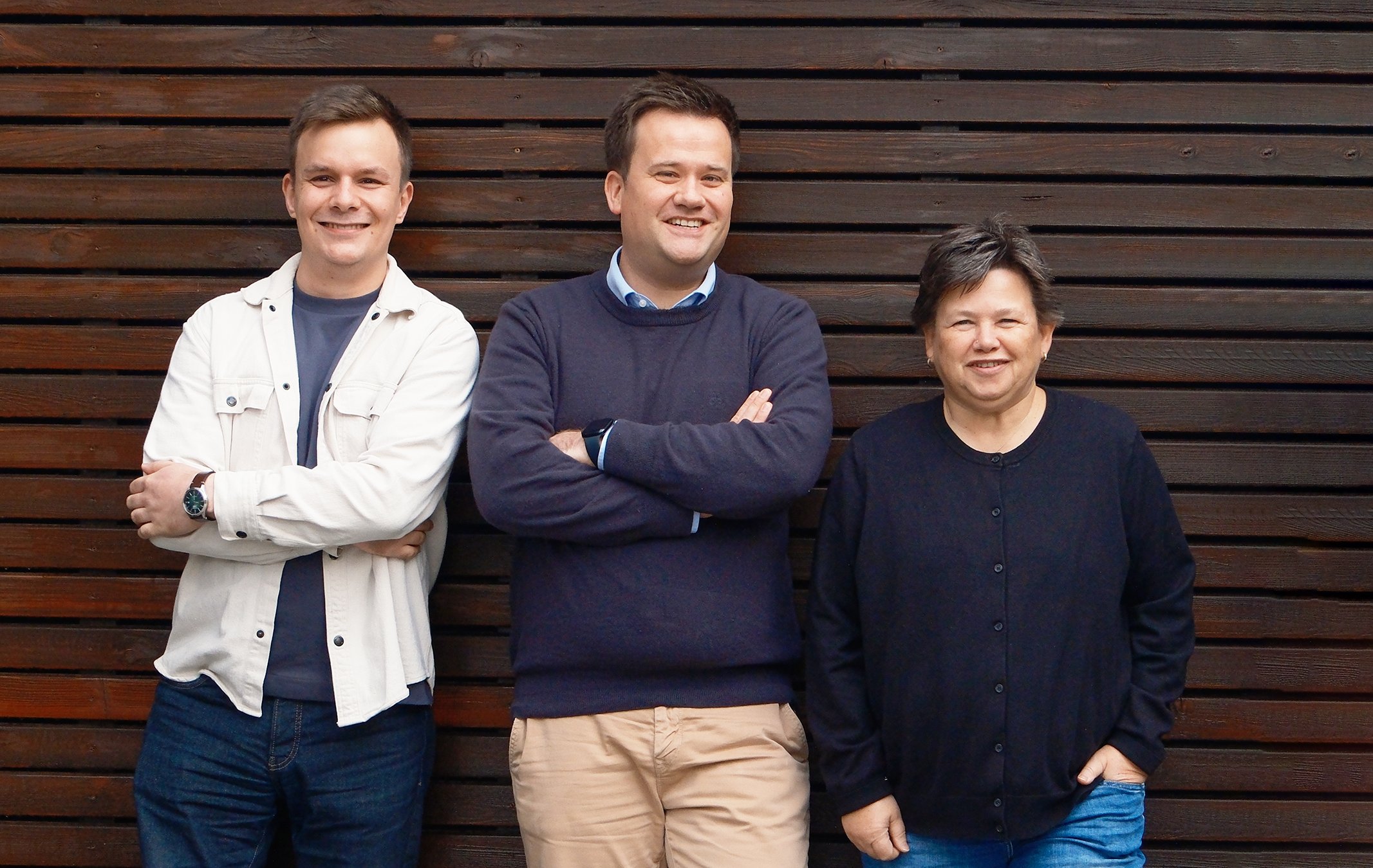 Zwei Männer und eine Frau stehen vor einer braunen Holzwand und schauen in die Kamera - TBE ingenieurbüro für Tragwerksplanung